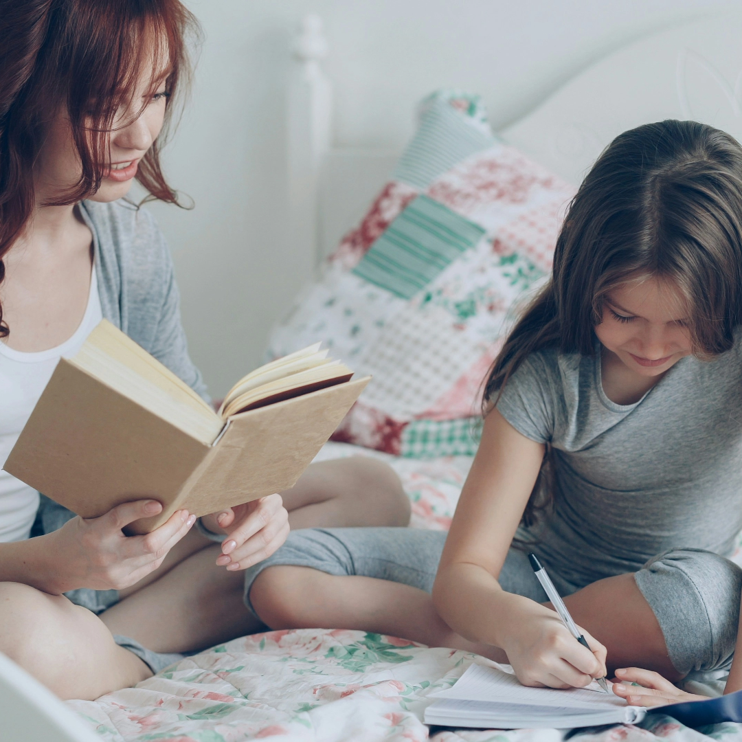 Two women sitting on a bed, one reading a book and the other writing in a notebook.